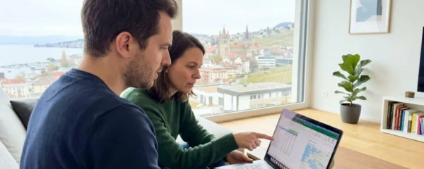 Un couple dans la trentaine consulte ensemble un ordinateur portable dans un salon contemporain lumineux avec vue floue sur les toits de Lausanne en arrière-plan